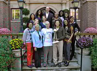 Three rows of people standing on steps outside the entrance to a brick mansion, with flowers and bushes on either side.